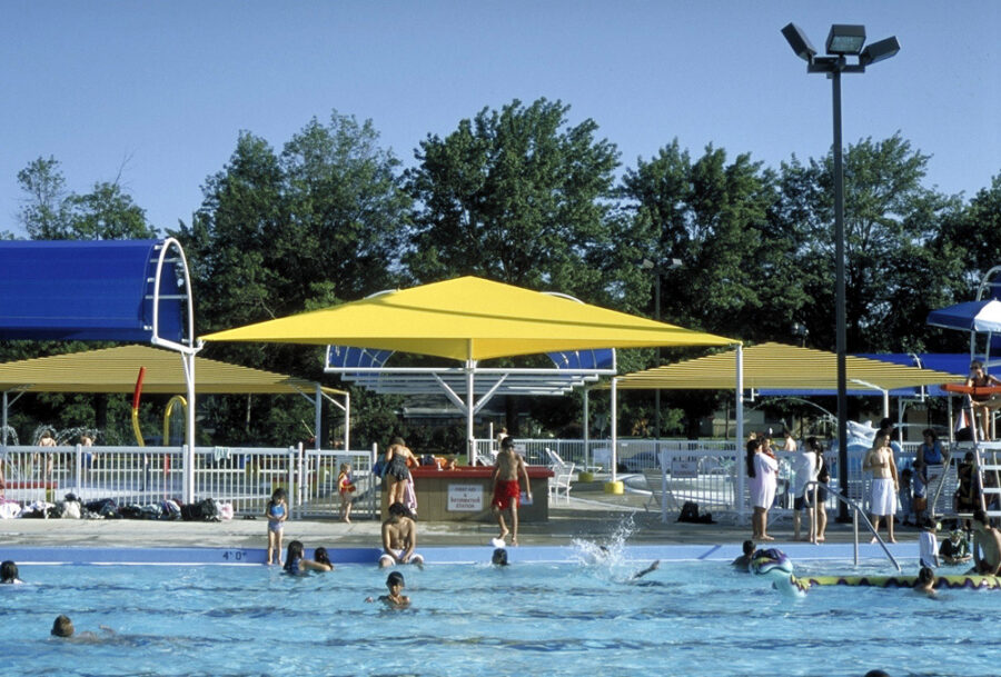 Outdoor swimming pool at Springdale Aquatic Complex in Arkansas featuring a large yellow shade canopy, covered seating areas and surrounding trees, designed by WDD Architects.